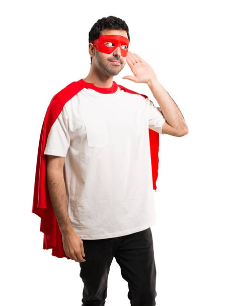 Superhelden-Mann mit Maske und rotem Umhang hört etwas, indem er die Hand auf das Ohr auf isoliertem weißem Hintergrund legt - Foto, Bild
