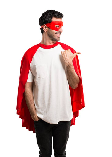 Bakarken bir ürün ya da bir fikir sunmak için maske ve kırmızı pelerin yan parmak ile işaret eden adamla süper kahraman ileri üzerinde gülümseyen izole beyaz arka plan - Fotoğraf, Görsel