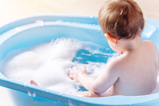 Χαριτωμένο μικρό παιδί αγόρι το πλύσιμο σε μπλε μπανιέρα στο μπάνιο. Το παιδί παίζει με νερό και σαπούνι αφρού. - Φωτογραφία, εικόνα