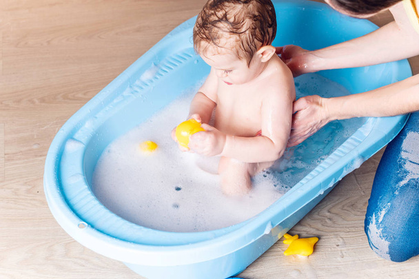 Mama prania mały chłopiec w niebieskim kąpieli w łazience. Dziecko bawi się żółta kaczka zabawka i bańki mydlane. - Zdjęcie, obraz