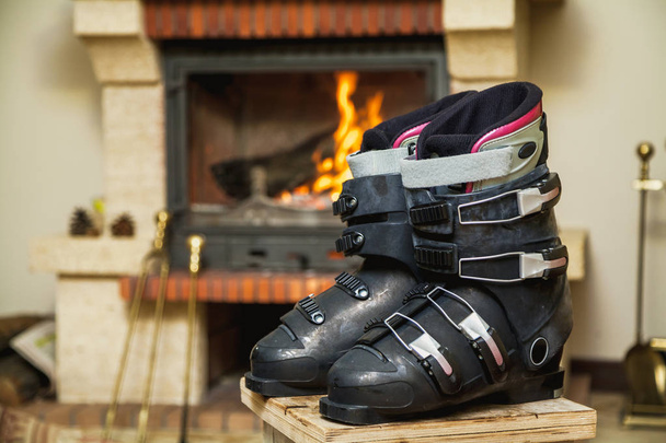 Drying shoes in front of the fireplace. Two ski boots stand on a stool in front of a burning fireplace in a bright roo - Photo, Image