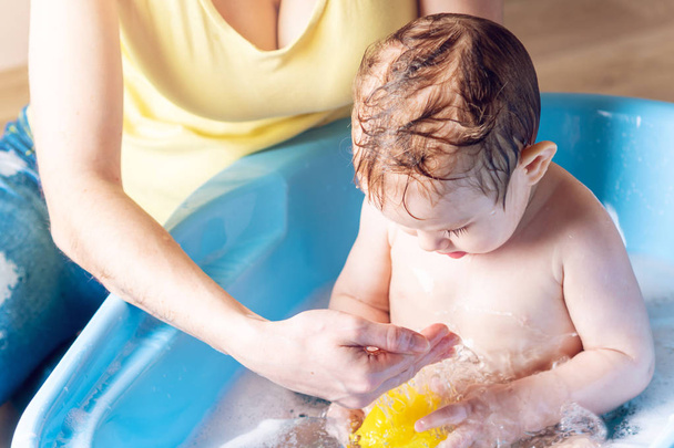 Máma mytí malého chlapce v modré lázni. Dítě je koupání s žluté kachny. Hygiena a vody léčba doma s dítětem - Fotografie, Obrázek