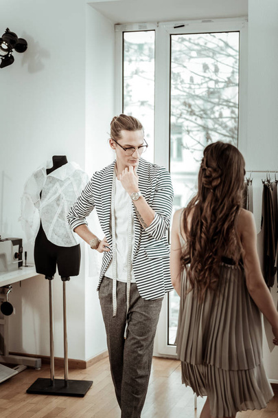 Last look. Long-haired fashion designer in eyeglasses looking thoughtful while looking at the model in a new dress - Φωτογραφία, εικόνα
