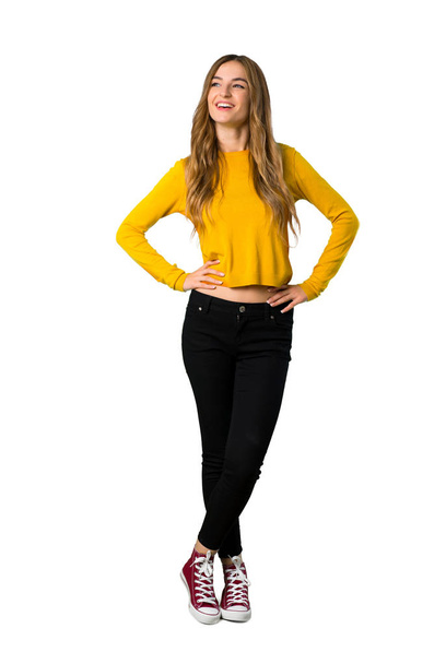 A full-length shot of a young girl with yellow sweater posing with arms at hip and laughing on isolated white background - Фото, изображение