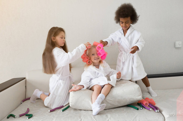 Little girls play with hair curlers and hairpins. Girls girlfriends in white coats braid curlers and hair clips each drg in her hair. - Foto, immagini