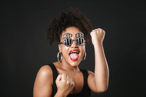 Portret van een mooie afro-Amerikaanse vrouw dragen van een zonnebril permanent geïsoleerd op zwarte achtergrond, vieren van succes - Foto, afbeelding