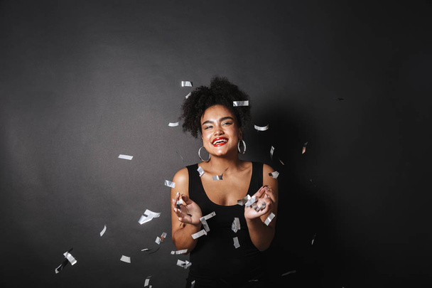 Photo of smiling afro american woman wearing black dress standing under falling confetti isolated over dark background - Фото, зображення