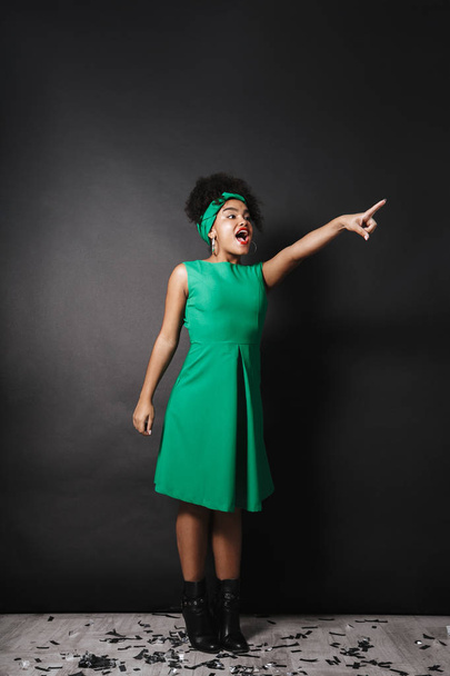 Full length photo of happy afro american lady wearing green dress pointing finger aside isolated over dark background - Fotó, kép