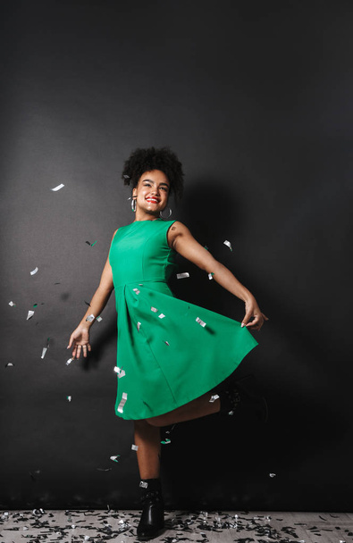 Full length portrait of a cheerful afro american woman wearing dress standing over black wall, posing under confetti shower - 写真・画像