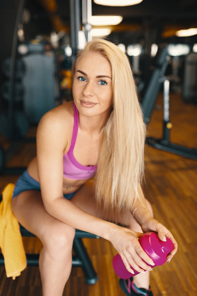 Mujer bonita con el pelo largo y rubio relajante en el gimnasio con una botella de agua rosa con equipo en el fondo y hermoso suelo de madera
. - Foto, imagen
