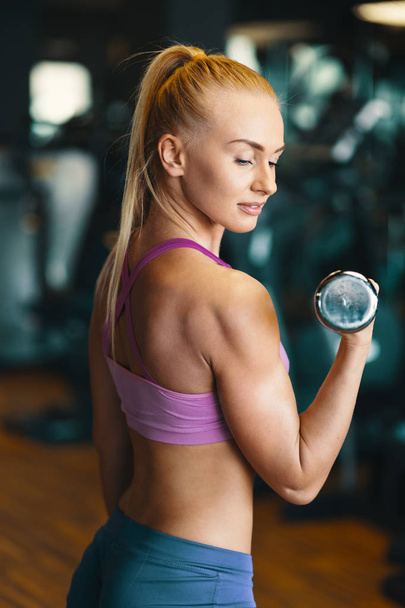 Young Caucasian woman doing exercise with dumbbells while training arms at the gym. Strength and motivation concept. Side view  - Zdjęcie, obraz
