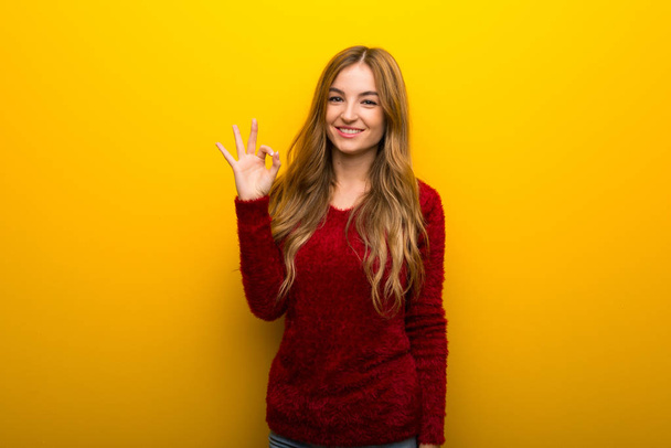 Νεαρό κορίτσι με ζωντανό κίτρινο φόντο δείχνει ένα ok σημάδι με τα δάχτυλα - Φωτογραφία, εικόνα