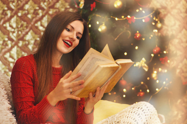 Nuori kaunis nainen punaisessa puserossa istuu neulotun huovan alla sohvalla ja lukee vanhaa kirjaa. Joulukuusi seppeleissä taustalla
. - Valokuva, kuva