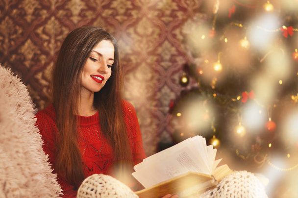 Kırmızı kazaklı genç güzel bir kadın örgü battaniye altında bir koltukta oturur ve eski bir kitap okur. Çelenk içinde belgili tanımlık geçmiş Noel ağacı. - Fotoğraf, Görsel
