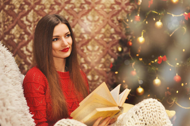 Une jeune belle femme en pull rouge s'assoit sous une couverture tricotée sur un canapé et lit un vieux livre. Arbre de Noël dans les guirlandes en arrière-plan
. - Photo, image