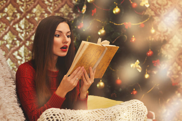 Een mooie vrouw die ook geïnteresseerd in de geschiedenis zit in een rode trui onder een gebreid deken op een bank en een oud boek leest. Kerstboom in de slingers op de achtergrond. - Foto, afbeelding