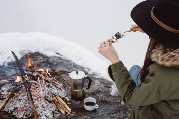 Girl eats fried marshmallows near campfire surrounded by snow against the near of the frozen lake. Concept adventure active vacations outdoor. Winter camp - Photo, image