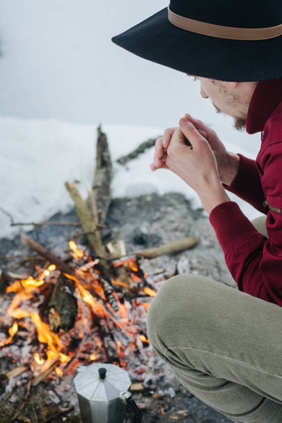 Winter camp. Tourist in hat cook coffee  and warms hands near a fire  an in winter time near of  frozen lake. Concept adventure active vacations outdoor.  - Fotoğraf, Görsel