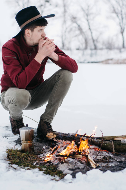 Winter camp. Tourist in hat cook coffee  and warms hands near a fire  an in winter time near of  frozen lake. Concept adventure active vacations outdoor.  - Fotografie, Obrázek