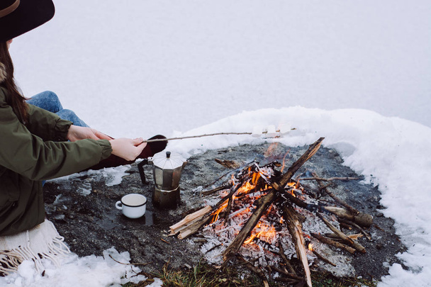 Girl roasting marshmallows over the campfire surrounded by snow against the near of the frozen lake. Concept adventure active vacations outdoor. Winter camp - Fotó, kép