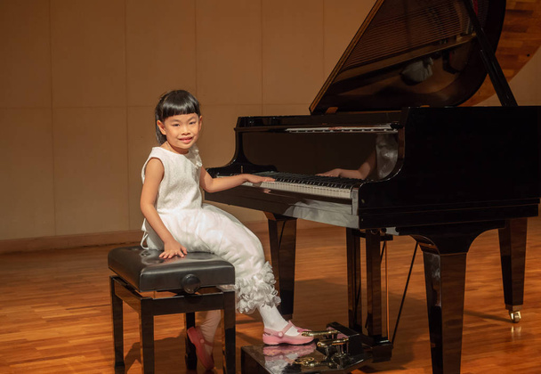 グランド ピアノのあるステージで白いドレスでアジアの少女。カメラを目線. - 写真・画像