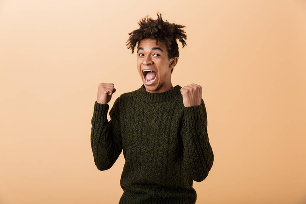 Portrait of an excited young african man dressed in sweater isolated over beige background, celebrating - Foto, afbeelding