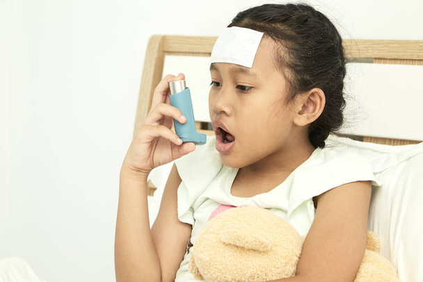 アジアの女の子がベッドの上に座っていると、broncodilator 吸入器を使用して喘息症状を和らげる - 写真・画像