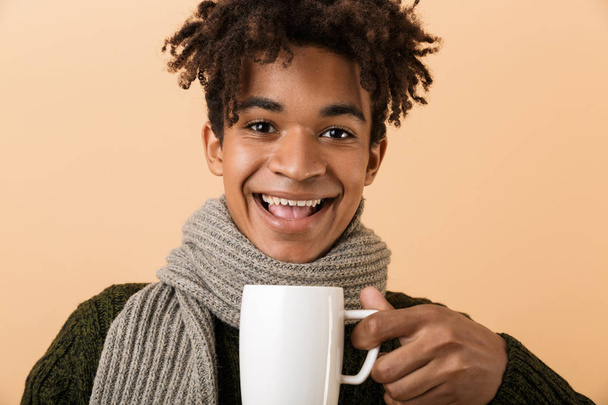 Close up portrait of a happy young african man dressed in autumn clothes isolated over beige background, holding a cup - Fotografie, Obrázek