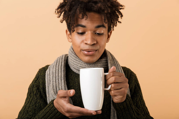 Close up portrait of a happy young african man dressed in autumn clothes isolated over beige background, holding a cup - Valokuva, kuva