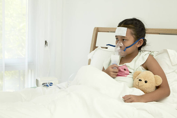 Azjatyckie dziewczyna siedzi na łóżku i za pomocą inhalatora broncodilator dla łagodzenia objawów astmy - Zdjęcie, obraz