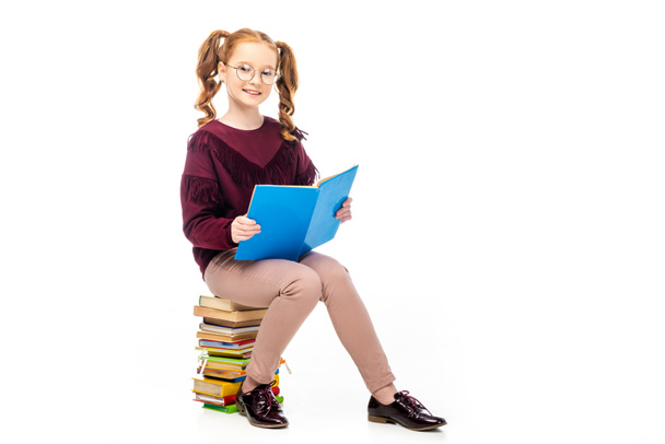 adorable schoolgirl in glasses sitting on books and smiling at camera isolated on white - Foto, imagen