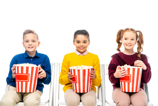 cute kids sitting on chairs, holding striped popcorn buckets and looking at camera isolated on white - Fotó, kép