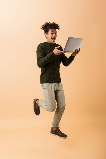Full length photo of happy african american guy wearing sweater walking with laptop isolated over beige background - Foto, Bild