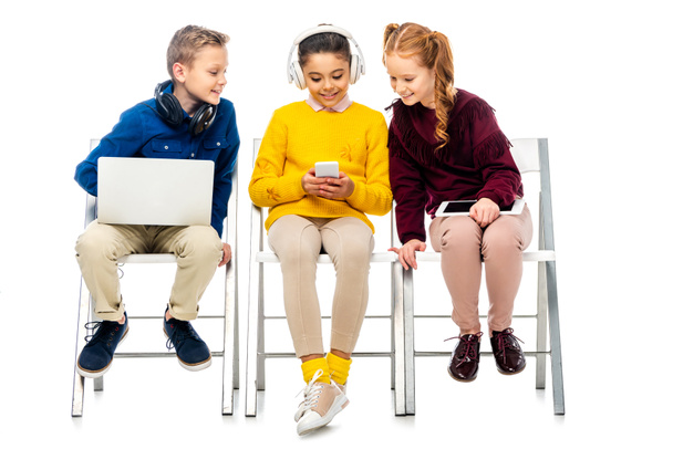 niños lindos sosteniendo dispositivos digitales y mirando la pantalla del teléfono inteligente de la colegiala linda con auriculares en la cabeza aislada en blanco
 - Foto, imagen
