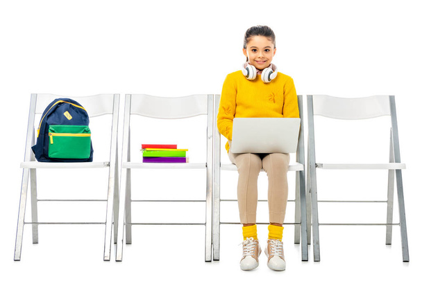 cute schoolgirl with headphones on neck sitting on chair, holding laptop and looking at camera isolated on white - Φωτογραφία, εικόνα