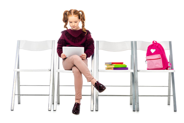 nettes Schulmädchen sitzt auf Stuhl und verwendet digitale Tablette isoliert auf weiß - Foto, Bild