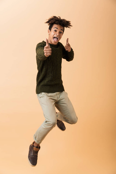 Full length photo of handsome african american guy wearing sweater fooling around and jumping isolated over beige background - Foto, afbeelding