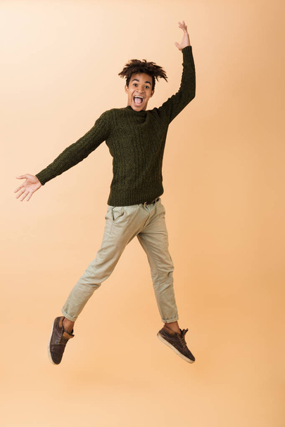 Volle lengte foto van blij Afro-Amerikaanse man dragen van de trui lachen en springen geïsoleerd op beige achtergrond - Foto, afbeelding