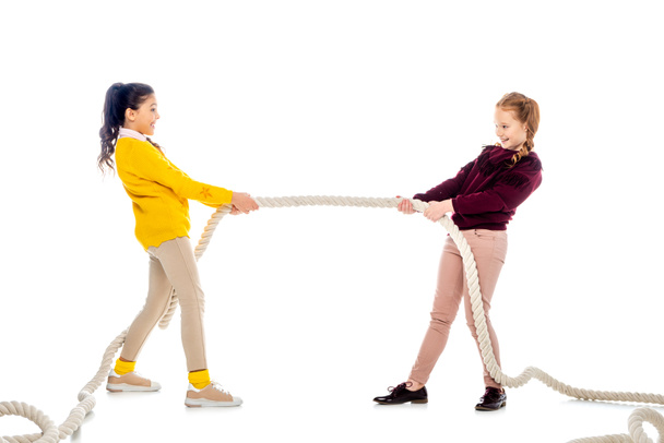 two cheerful schoolgirls pulling rope and looking at each other isolated on white - Foto, Imagem