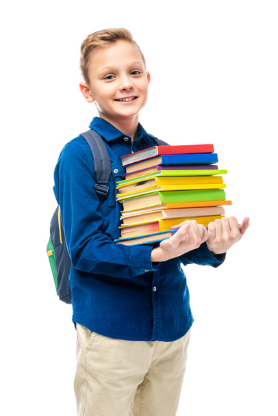 menino bonito com mochila segurando pilha de livros e olhando para a câmera isolada no branco
 - Foto, Imagem