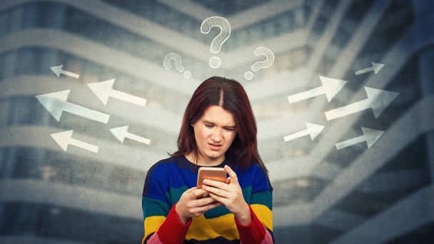 Woman looking at smartphone with puzzled face confused and frustrated has to choose arrow direction, left or right side, interrogation marks above head. Difficult decision concept, correct choice. - Photo, Image