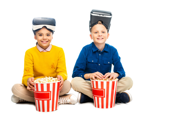 smiling kids with virtual reality headsets on heads holding striped popcorn buckets and looking at camera isolated on white - Fotografie, Obrázek