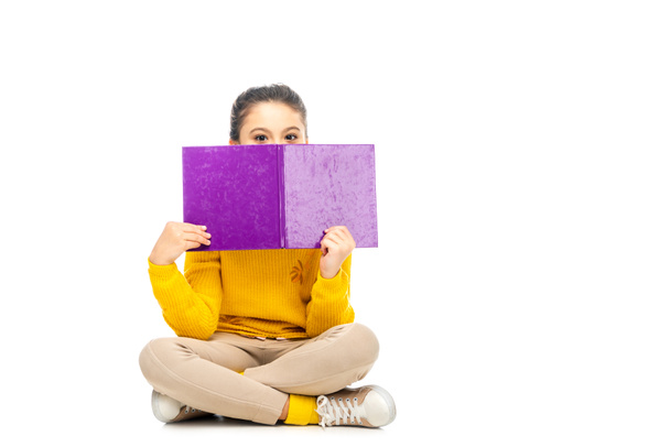 schoolgirl sitting and looking over top of purple book at camera isolated on white - Φωτογραφία, εικόνα