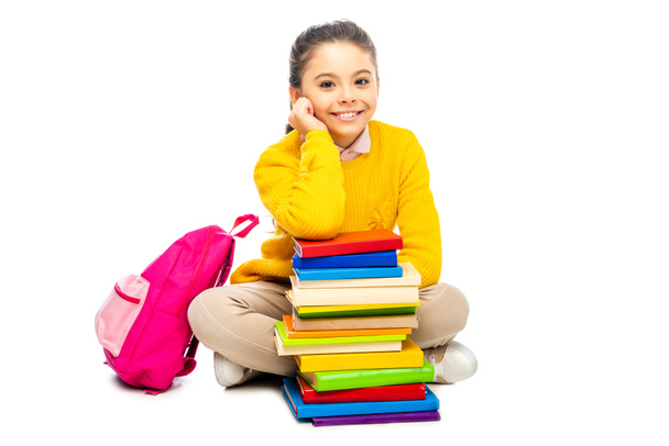 cute schoolgirl sitting near stack of books and pink backpack and looking at camera isolated on white - Foto, Bild