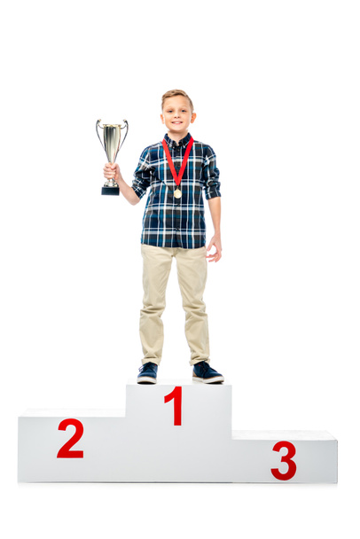 usměvavý chlapec stojící na podstavci, vítěz, držel trophy pohár a při pohledu na fotoaparát izolované na bílém - Fotografie, Obrázek