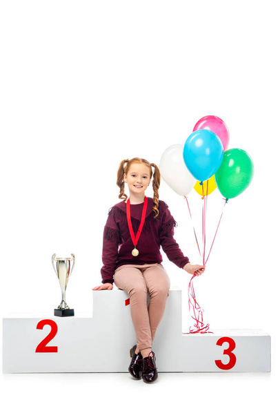 smiling schoolgirl sitting on winner podium near trophy cup, holding colorful balloons and looking at camera isolated on white - Photo, Image