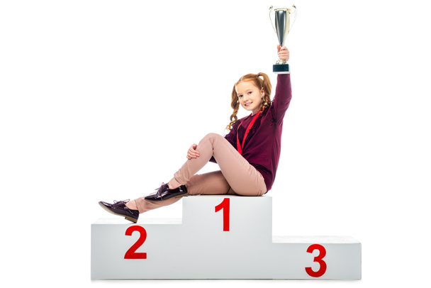 colegiala sonriente sentada en el podio ganador, sosteniendo la copa de trofeo en la mano levantada y mirando a la cámara aislada en blanco
 - Foto, Imagen