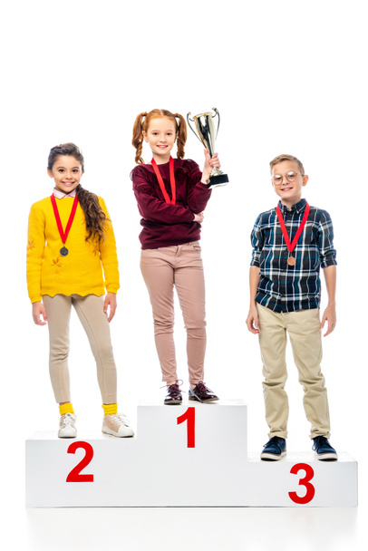 smiling preteen kids with medals and trophy cup standing on winner pedestal, smiling and looking at camera isolated on white - Photo, Image
