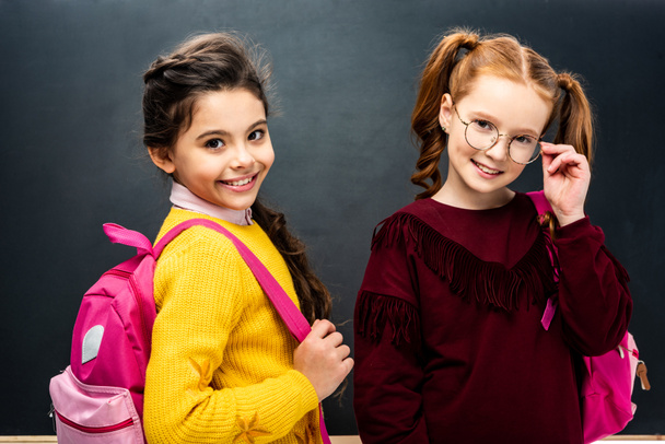 smiling schoolgirls with pink backpacks looking at camera on black background - Фото, изображение
