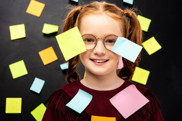 cute schoolgirl in glasses with multicolored stickers smiling and looking at camera on black background - Fotografie, Obrázek